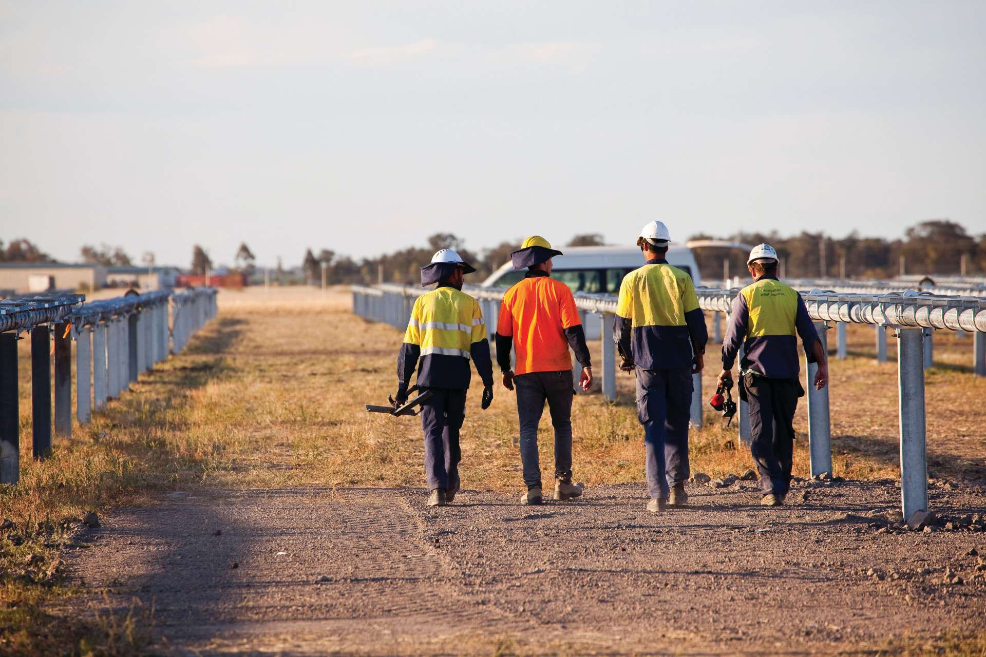 Photo of four workers in PPE workwear carrying various tools walking through a rural electricity generation worksite.